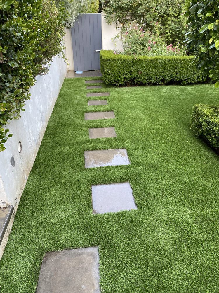 A stone-stepped path leads through a lush green artificial lawn toward a colonial-style building with a white balcony.