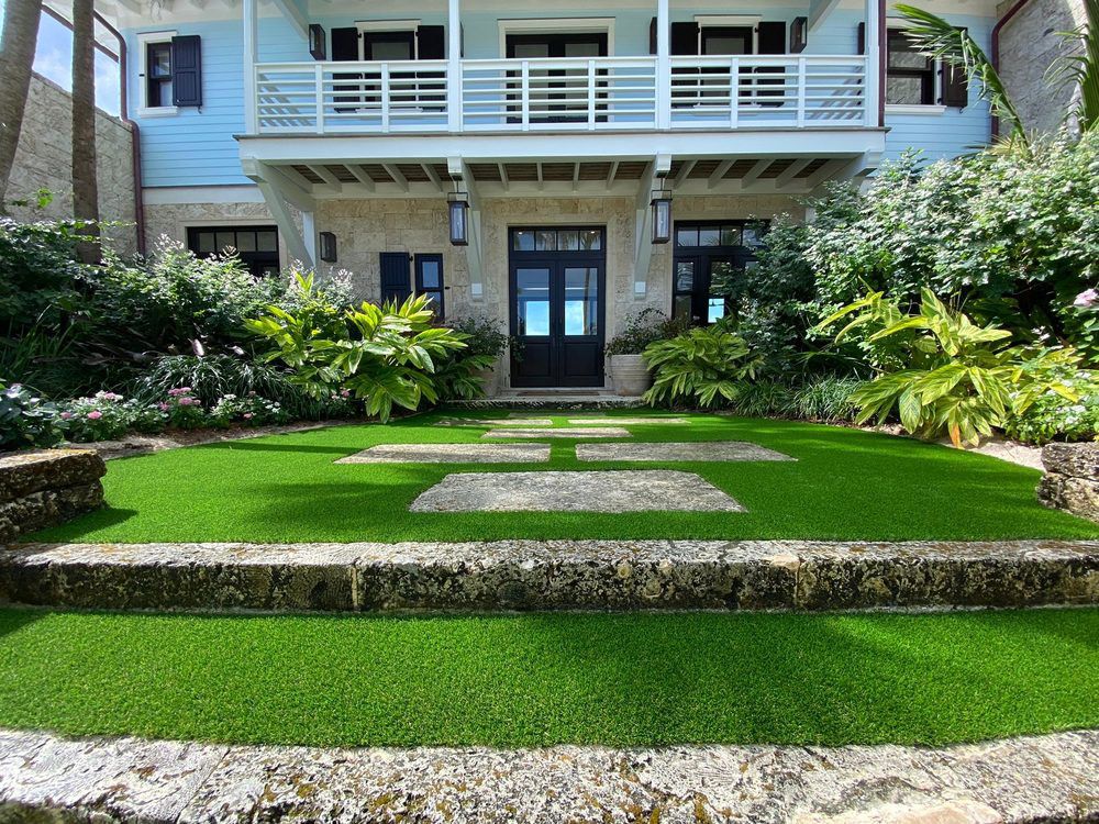 A tiered artificial lawn with stone pavers leads to a two-story blue and beige house with a balcony and lush landscaping.