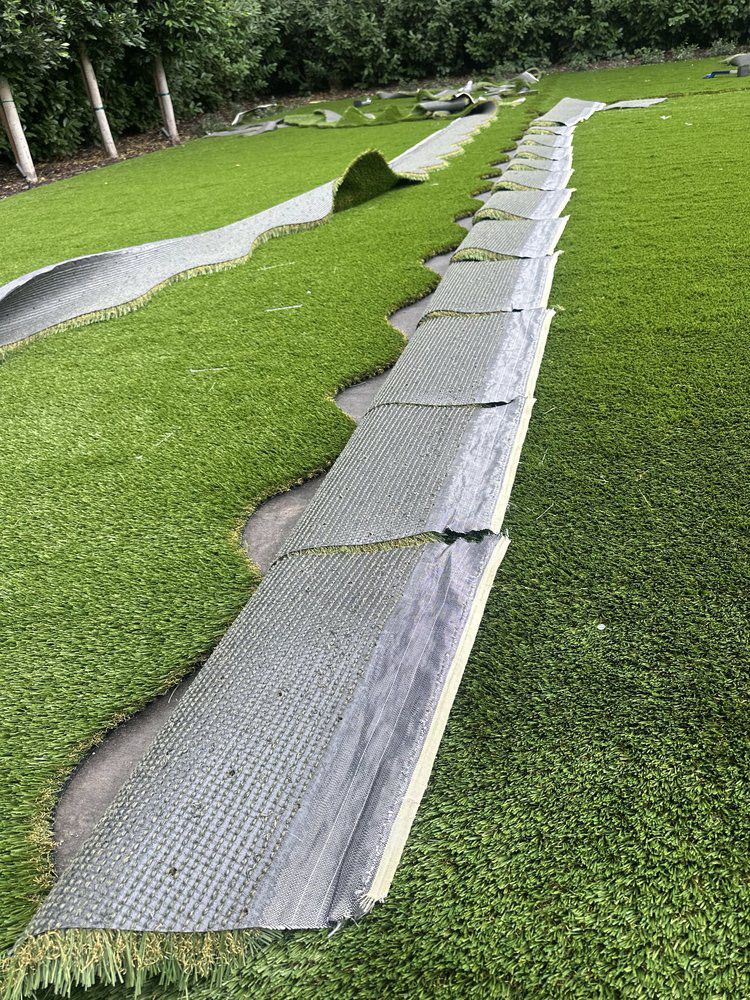 Strips of synthetic turf lie on a green lawn, revealing a grey, grid-patterned backing along the seams.