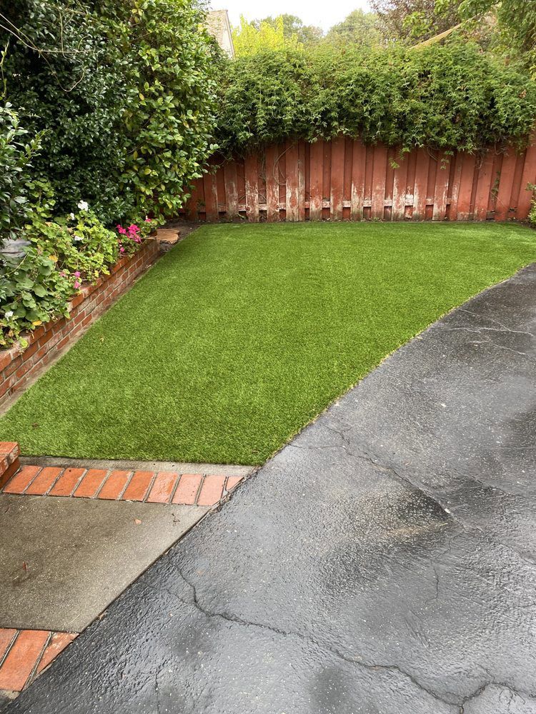 A green lawn bordered by a brick edge and a dark asphalt driveway, set against a red fence and lush green hedges.