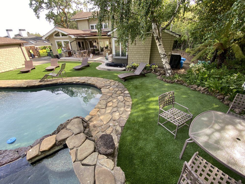 A backyard scene with a rectangular swimming pool, raised hot tub, modern pergola, and lush green synthetic grass lawn.