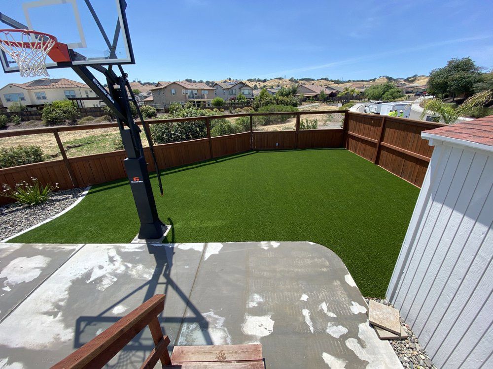 A residential backyard featuring a basketball hoop on a concrete patio next to a patch of green artificial turf and fence.