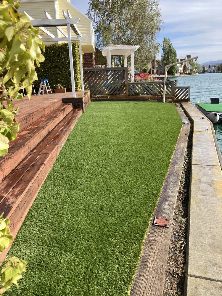 A rectangular patch of artificial grass sits between a wooden deck and a stone dock edge beside a calm, blue lake.