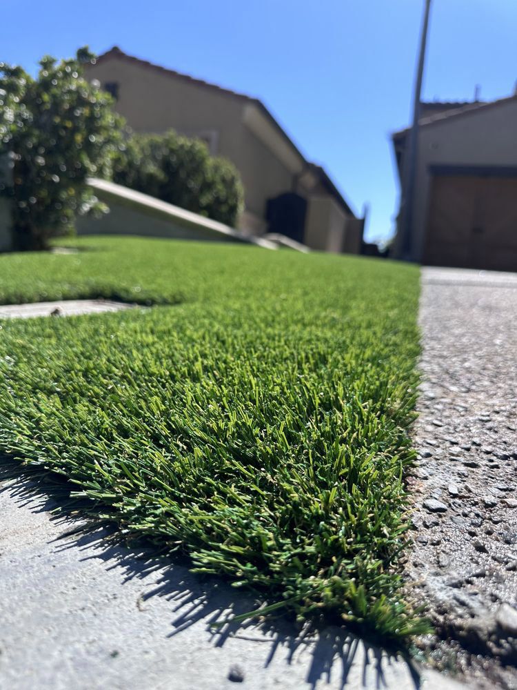 A low-angle view of vibrant green artificial grass bordering a concrete sidewalk, with residential houses in the background.