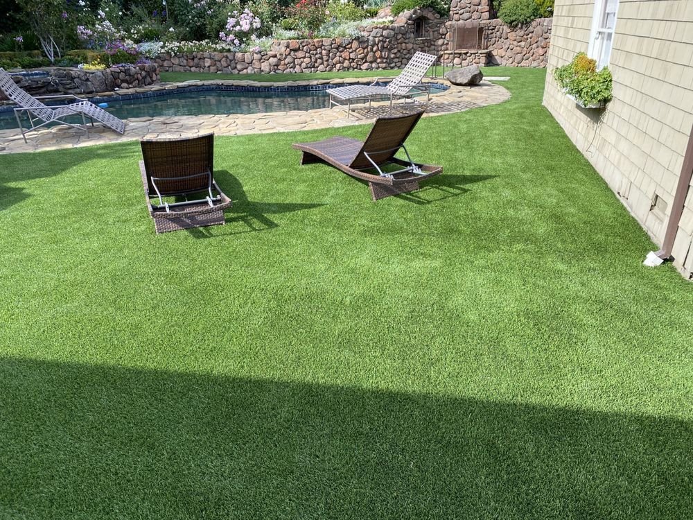 Two lounge chairs sit on a green artificial lawn next to a backyard pool and a stone wall.