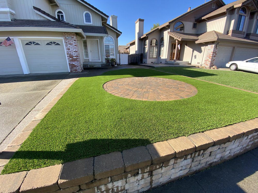 A residential front yard featuring a circular paver patio surrounded by green synthetic turf and a low stone retaining wall.