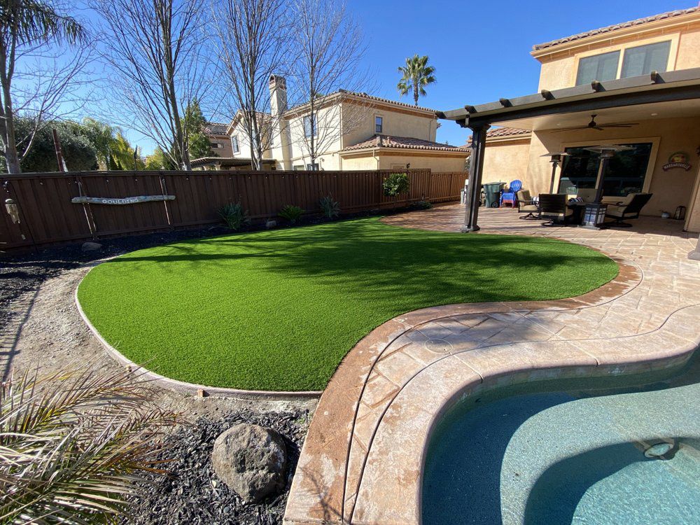 Backyard with a curved artificial grass lawn, a stone-bordered pool, and a covered patio area under a sunny blue sky.