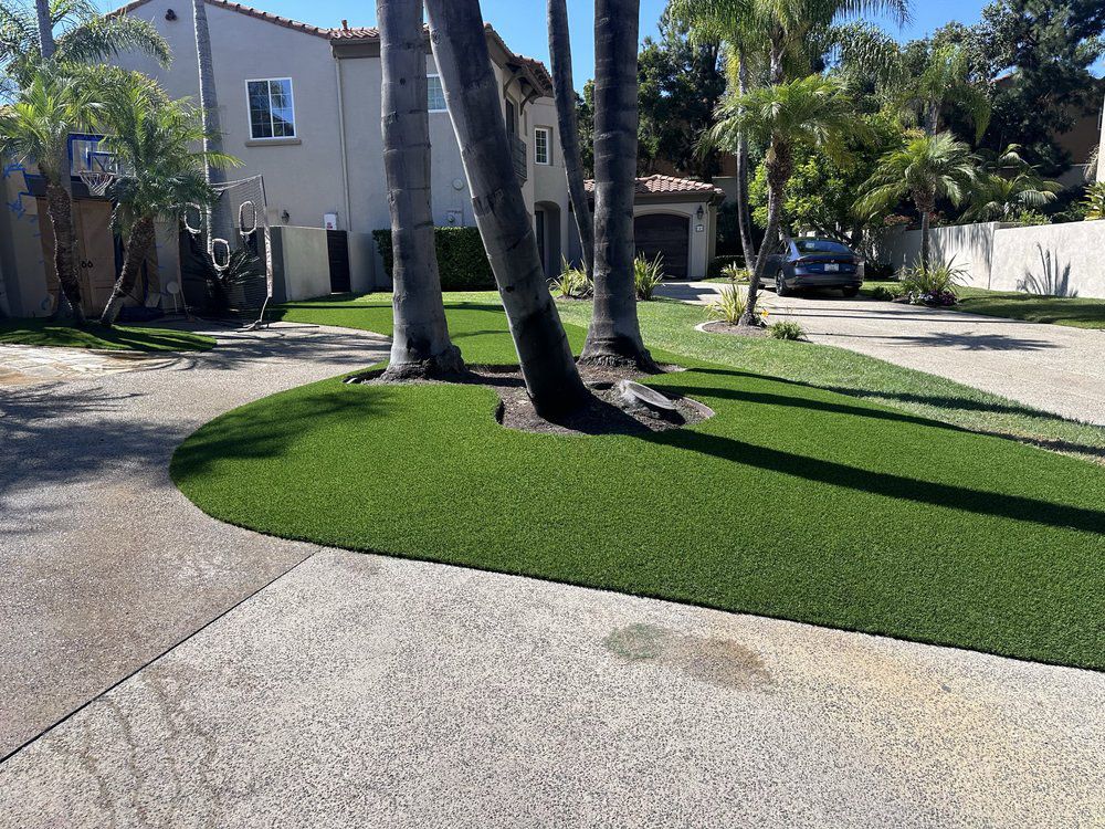A residential front yard features a manicured patch of artificial green grass surrounding two palm trees near a driveway.