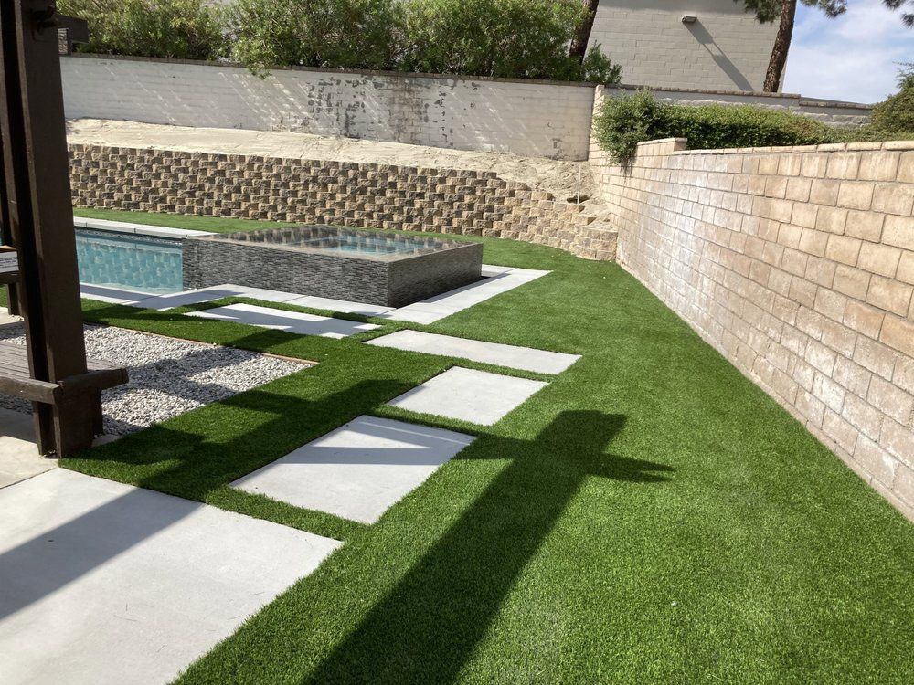 A backyard with artificial turf, concrete stepping stones, a swimming pool, and stone retaining walls under a sunny sky.