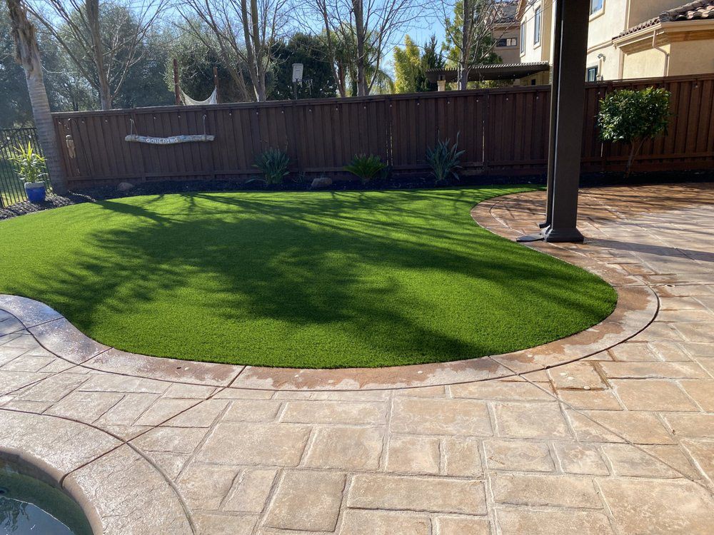 A patch of green artificial grass surrounded by a textured, tan stone patio, with a wooden fence in the background.