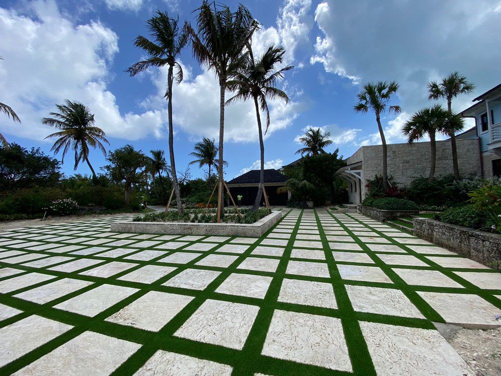A patio with white square pavers separated by grass strips, featuring palm trees and a building under a cloudy blue sky.
