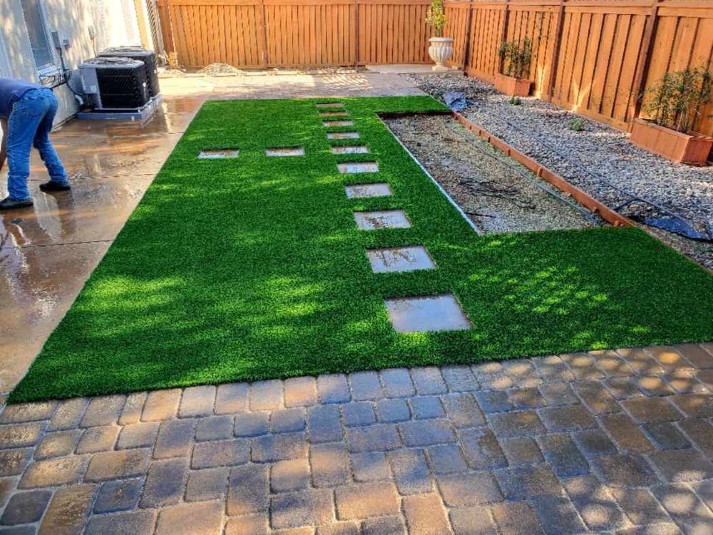A person installing artificial turf in a backyard with a stone paver path and a gravel area along a wood fence.