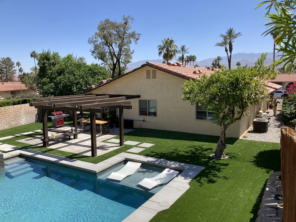 A backyard with a swimming pool, a tanning ledge with lounge chairs, a wooden pergola, and a house under a clear blue sky.