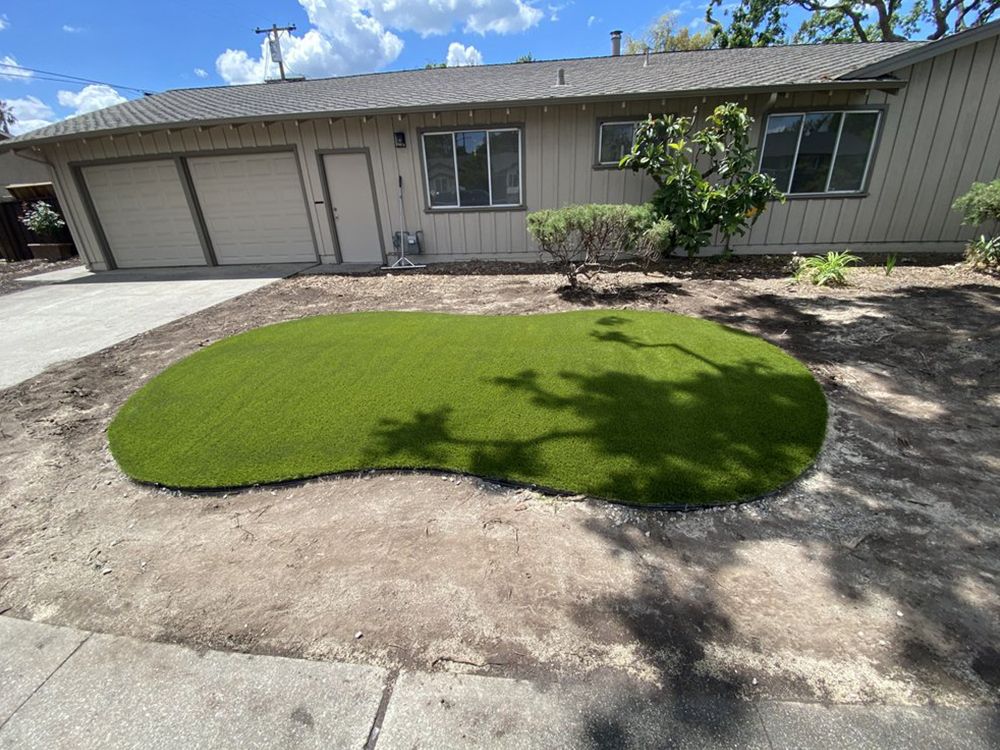 An oval patch of bright green artificial turf installed in a front yard in front of a tan house with a garage.