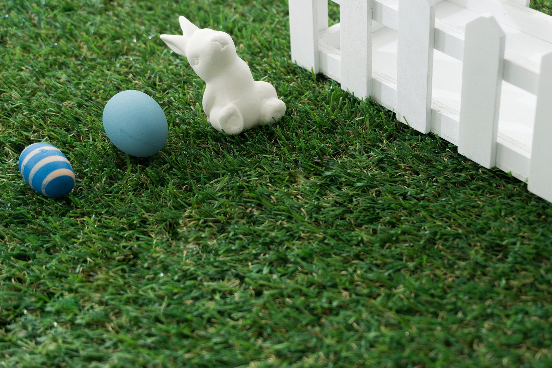 A white rabbit figurine, a plain blue egg, and a blue-and-white striped egg sit on green grass next to a white fence.