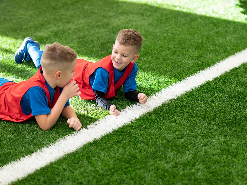 Two young children in red vests and blue shirts lie on green turf near a white line, one smiling.