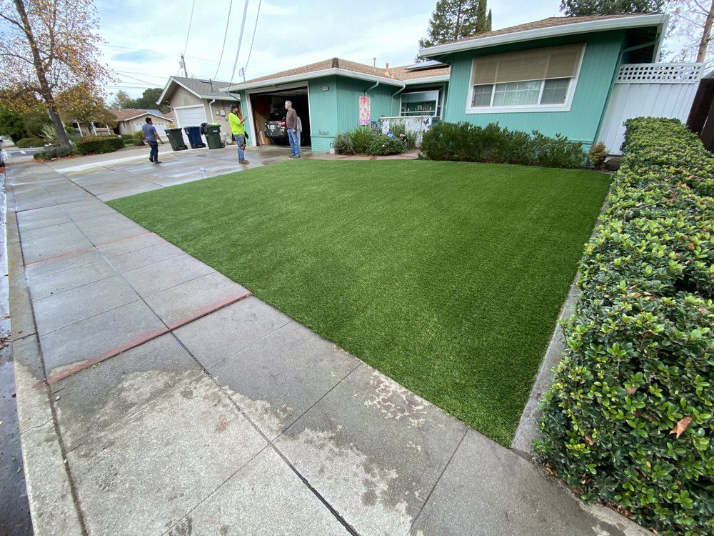 A green, manicured artificial lawn in front of a teal single-story house with people working in the open garage.
