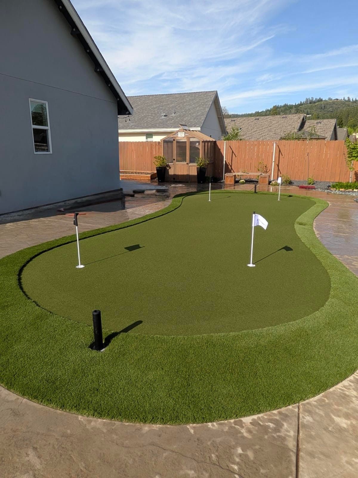 A backyard putting green with several holes and flags set in a residential area with a house and gazebo nearby.