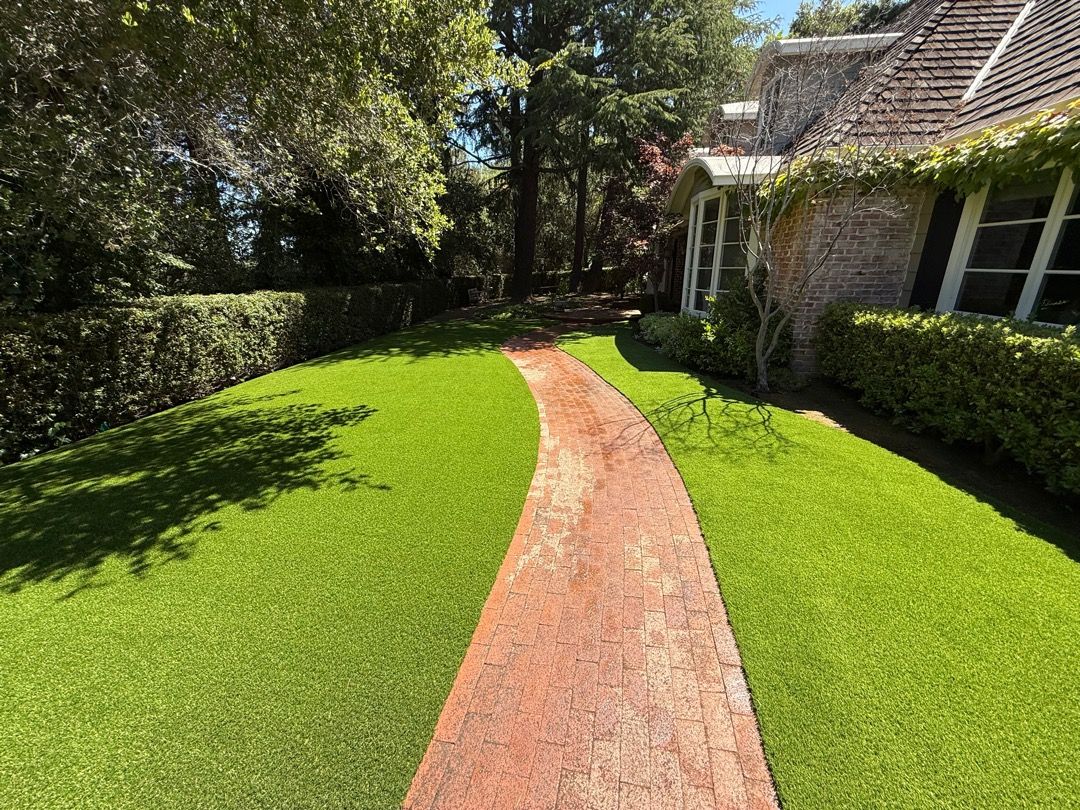 A brick walkway curves through a vibrant green lawn towards the side of a brick house with large windows.