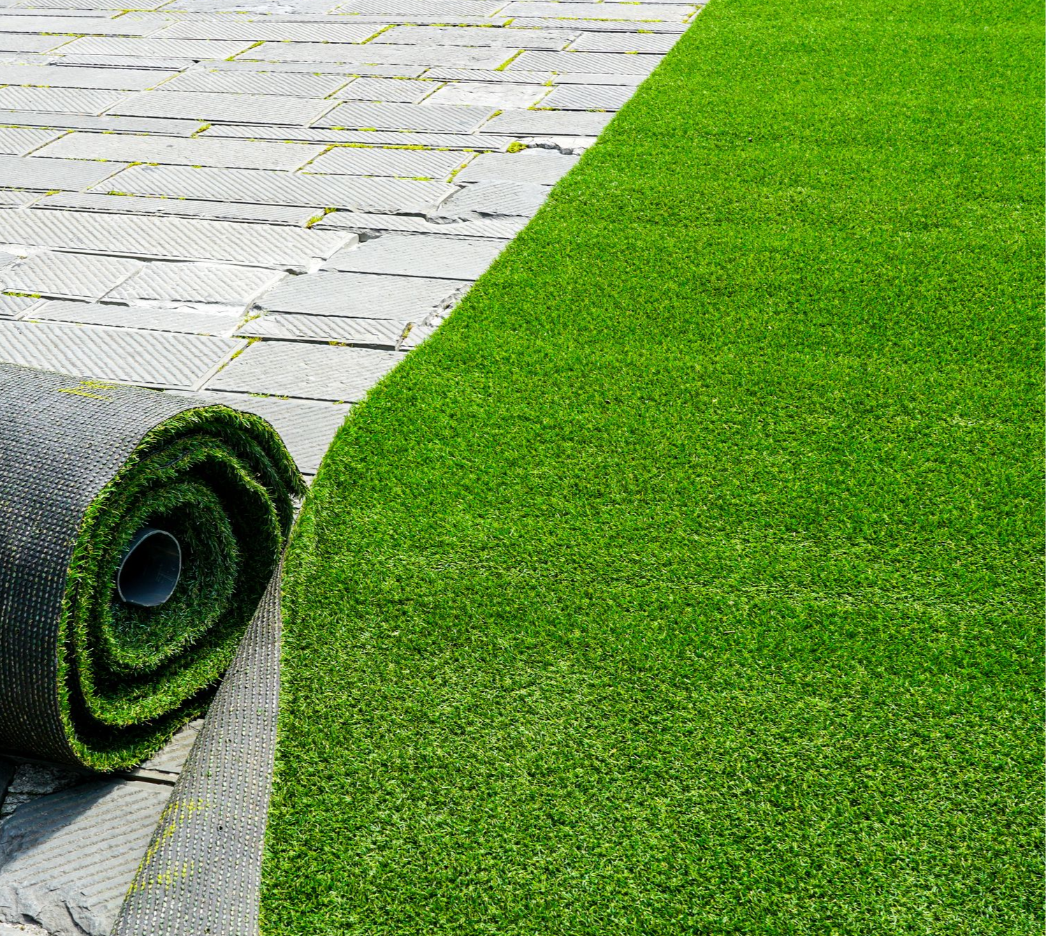 A roll of bright green artificial turf being unrolled onto a grey paved surface.