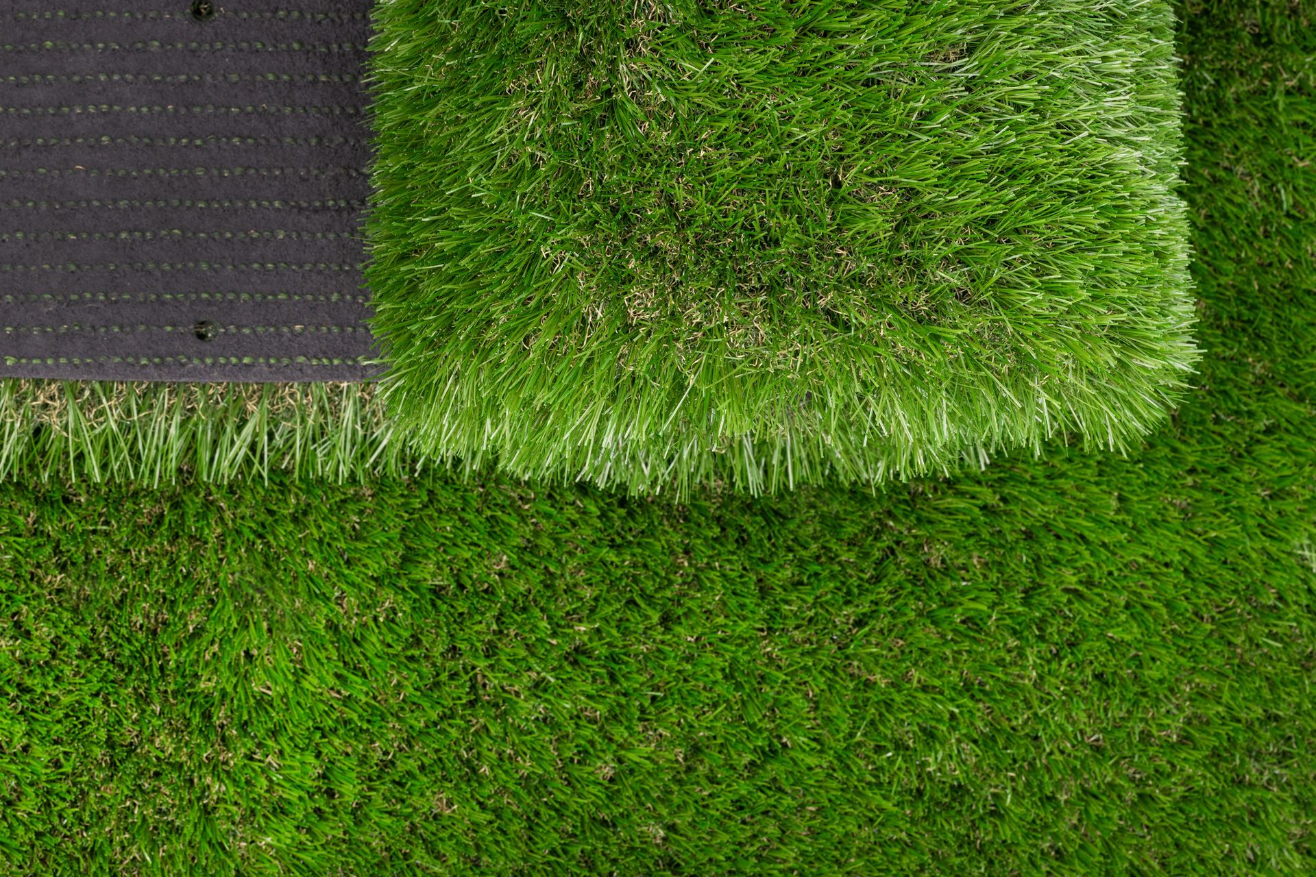 A patch of vibrant green artificial grass resting on top of a larger section of the same turf with a visible black backing.