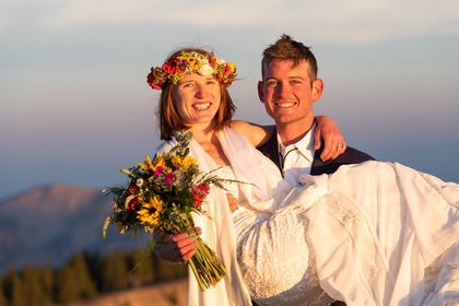 Smiling couple in wedding attire outdoors at sunset, with a floral crown and bouquet