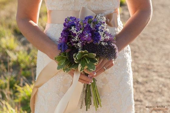 Bridal bouquet of purple and white flowers held at a bride’s waist in a sunlit outdoor setting
