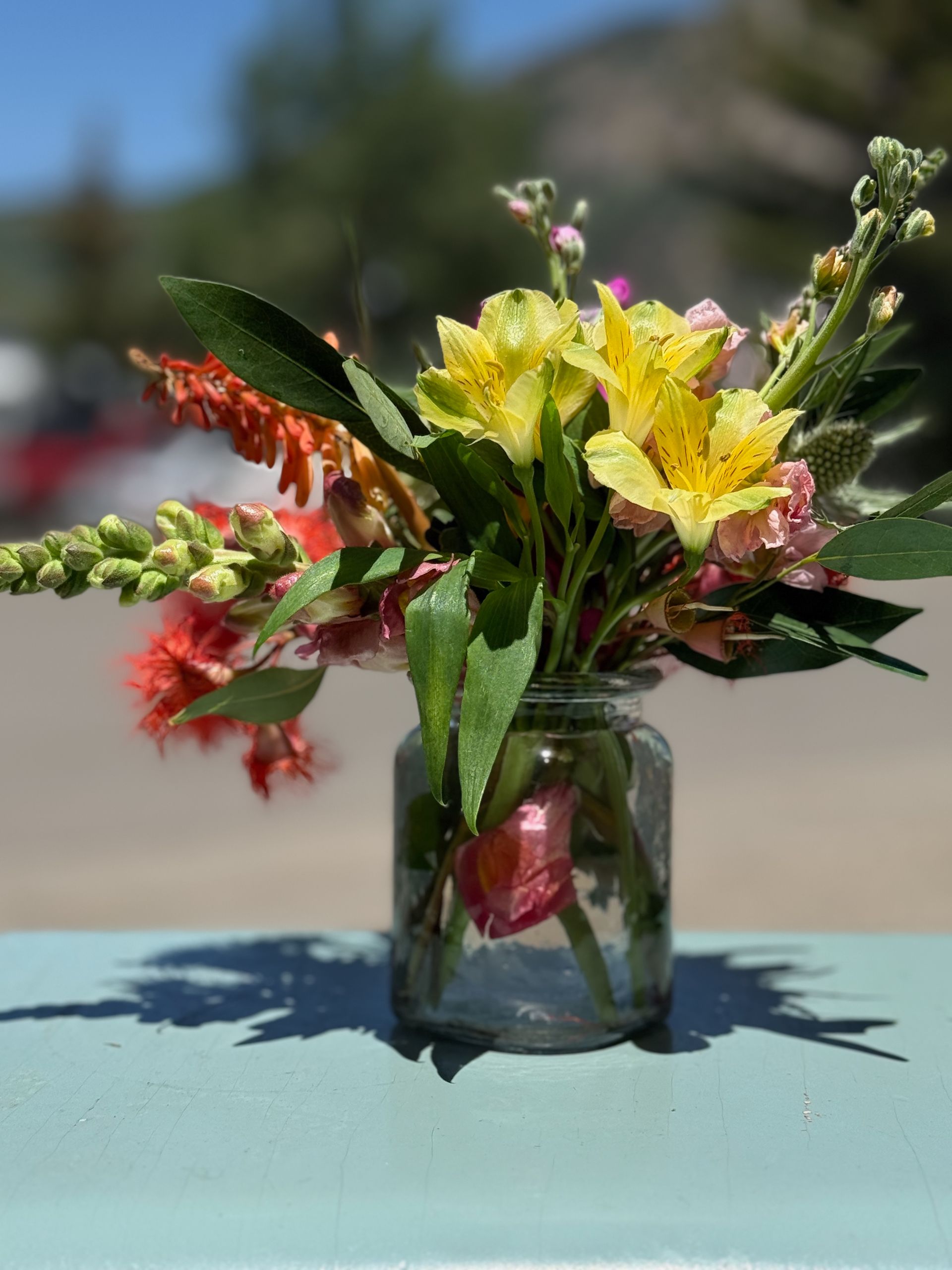 Small bouquet of colorful wildflowers in a glass jar on a blue surface outdoors