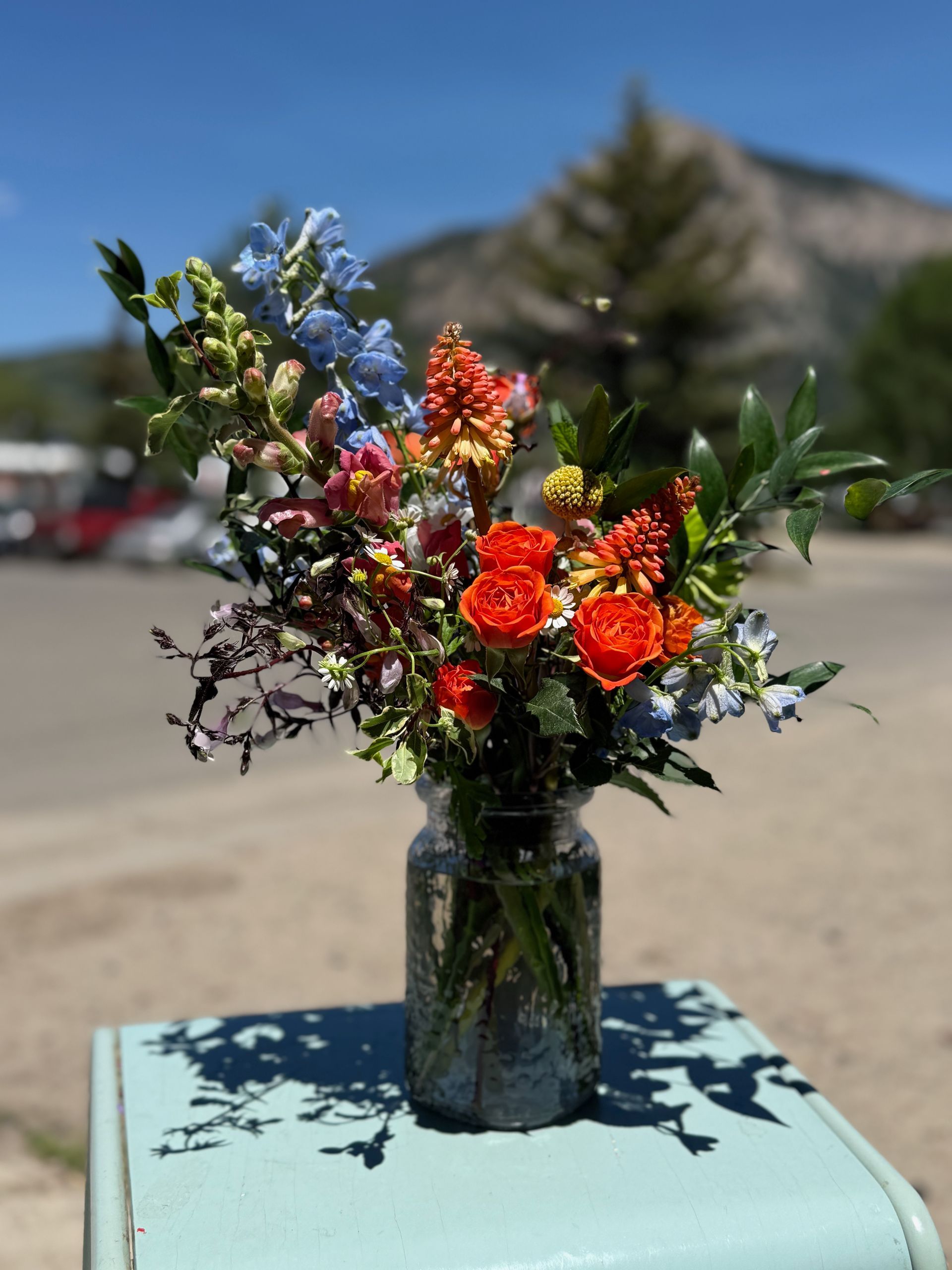 Wildflower bouquet in a dark vase on a blue table outdoors, with mountains in the background