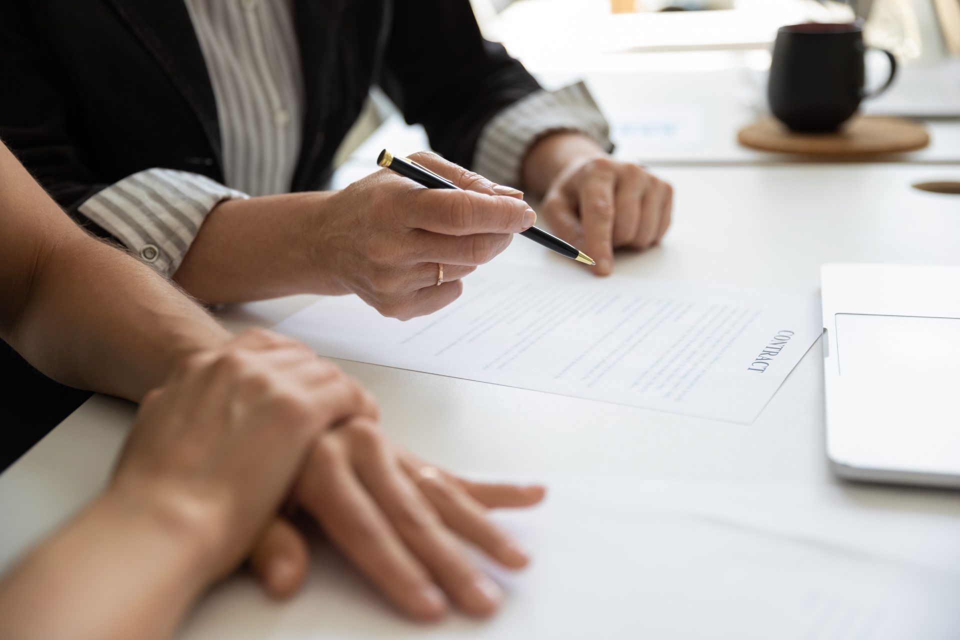 People signing a document at a white desk with a laptop and coffee mug nearby