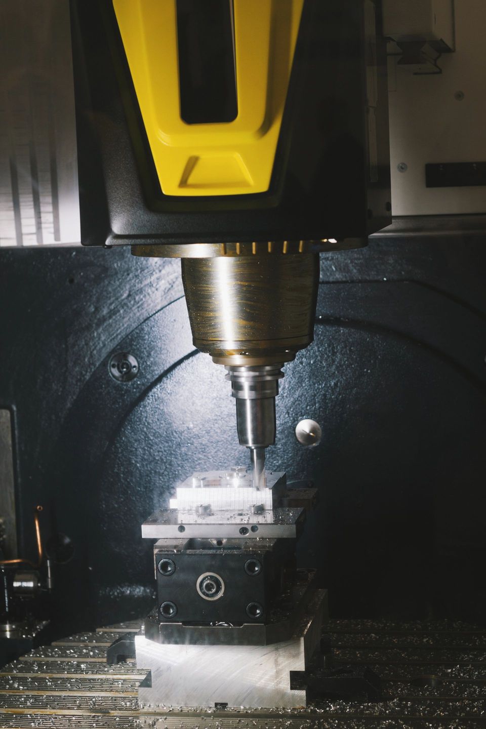 Una macchina a controllo numerico con una testa dai dettagli gialli fresa un pezzo metallico fissato in una morsa nera all'interno di un involucro industriale.