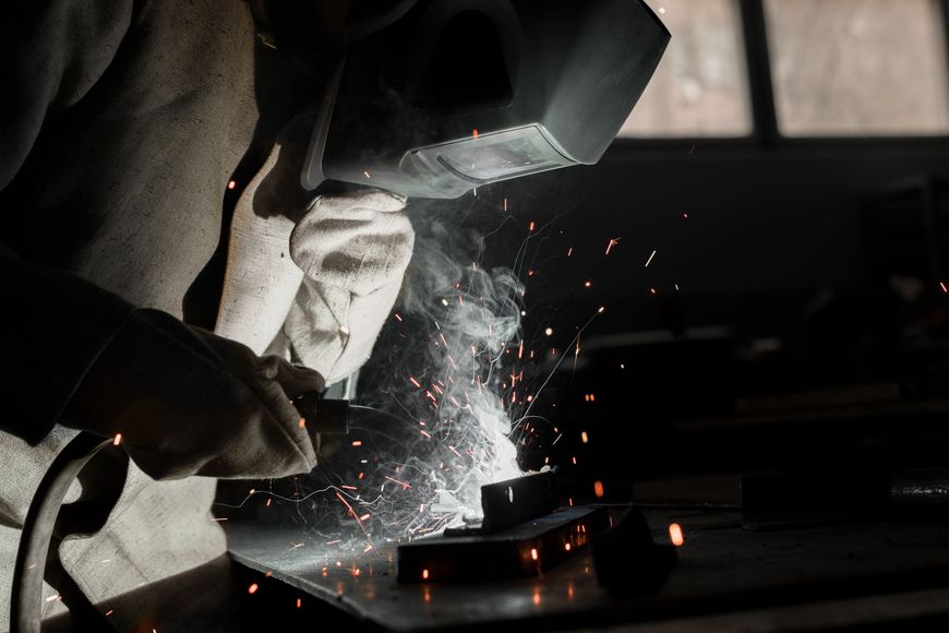 Un saldatore con indumenti protettivi lavora sul metallo, creando scintille luminose e fumo in un'officina scarsamente illuminata.