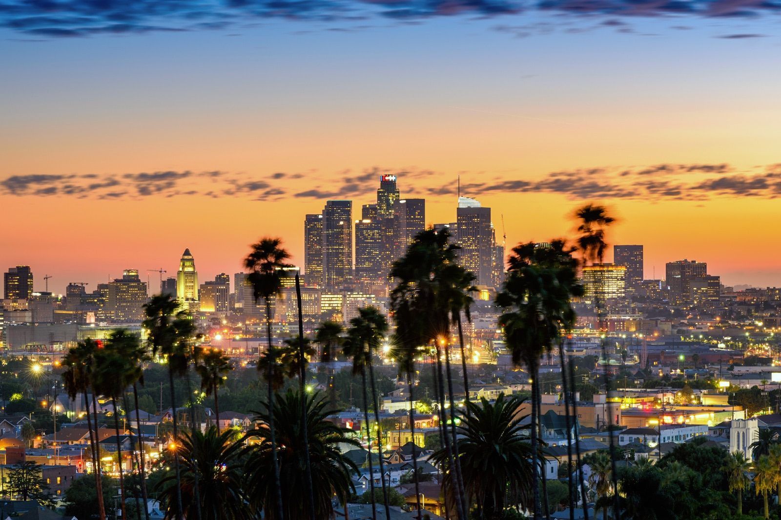 City skyline at sunset with palm trees and glowing lights under a colorful dusk sky