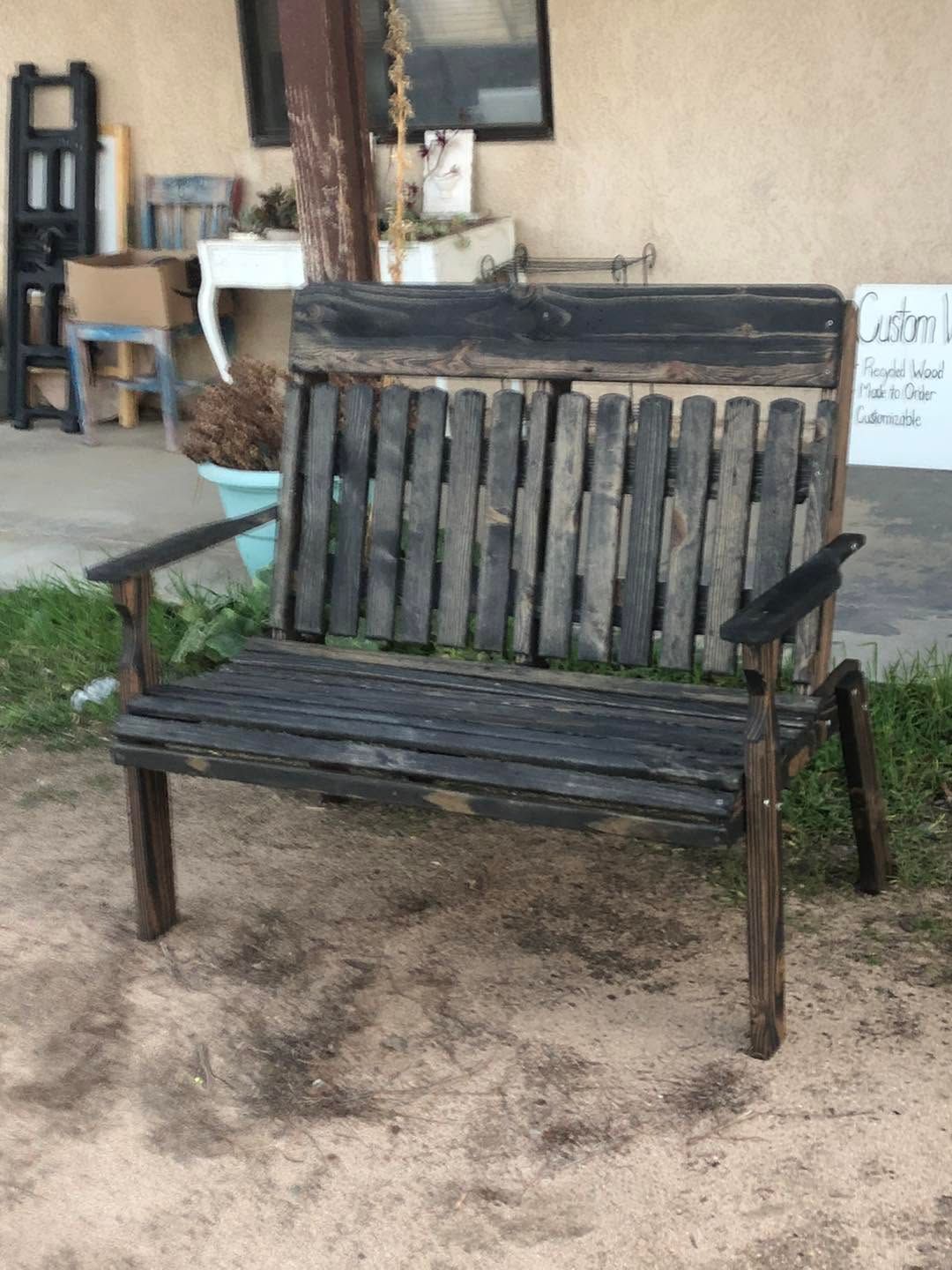 Rustic wooden bench with slatted back and seat, sitting on dirt outdoors near a building.