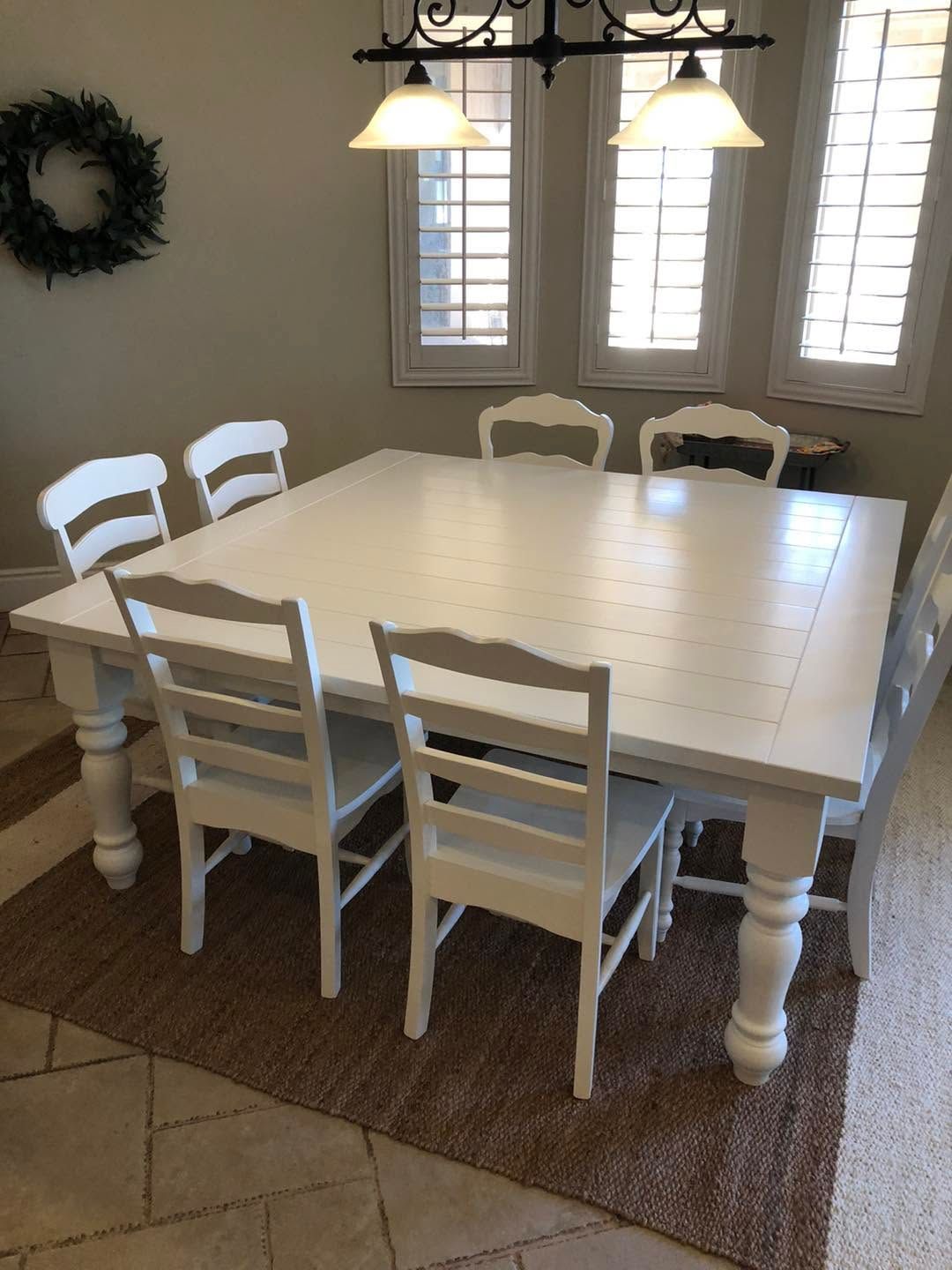 White dining table with six chairs on a woven rug in a bright room with windows and a wreath on the wall