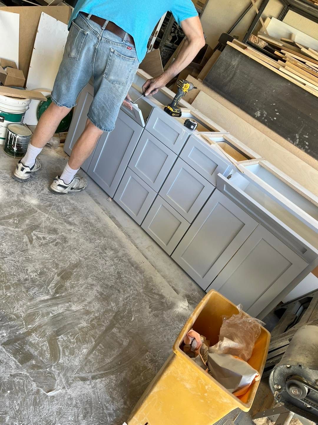 Person in a blue shirt and shorts working at a countertop in a workshop or garage with cabinets and tools.