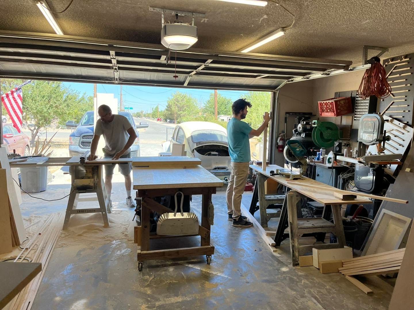 Two people working in a garage workshop with a table saw and tools, open door showing parked cars outside