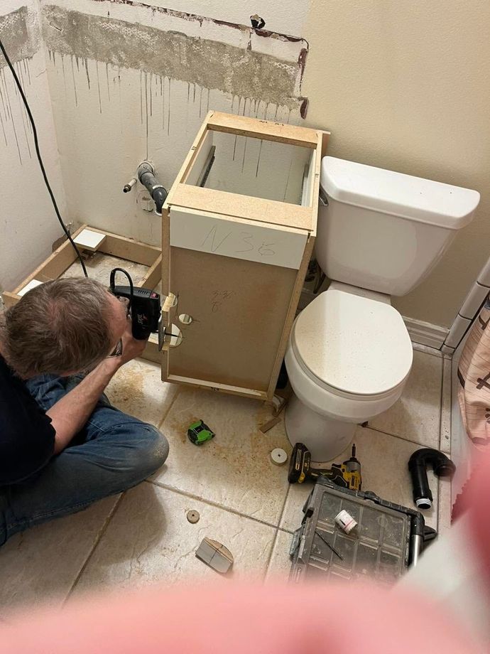 Person kneeling and repairing a bathroom vanity beside a toilet, with tools and exposed plumbing connections.
