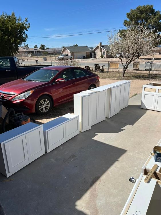 White cabinet doors and panels lined up on a driveway beside a red car under a blue sky