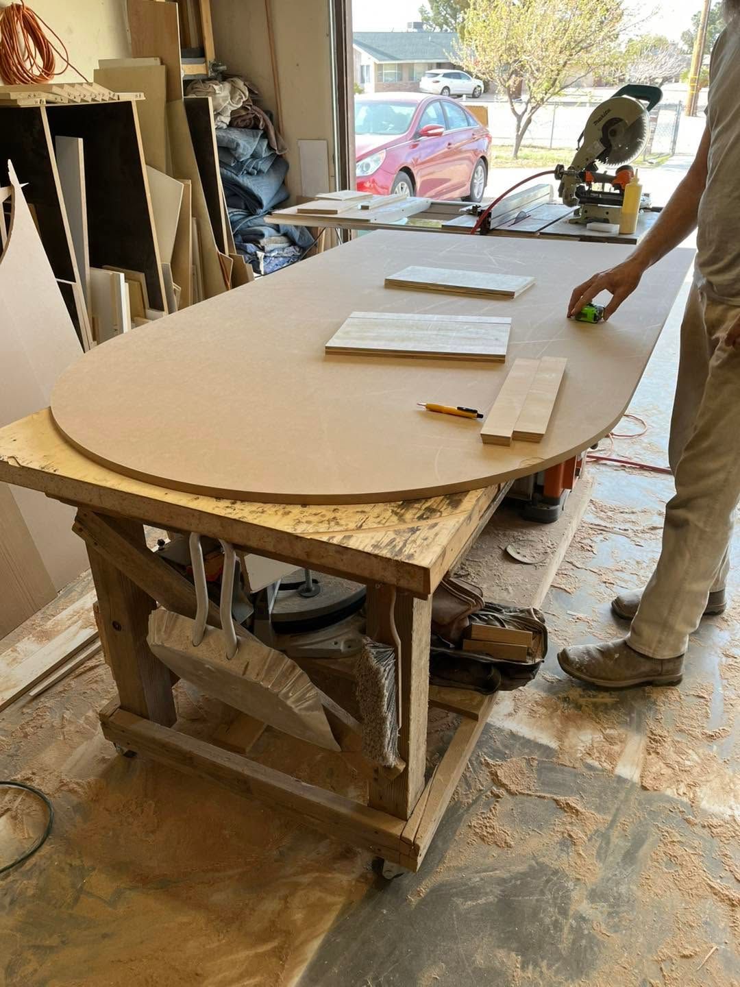 Person sanding a large curved wooden panel in a workshop by an open garage door.