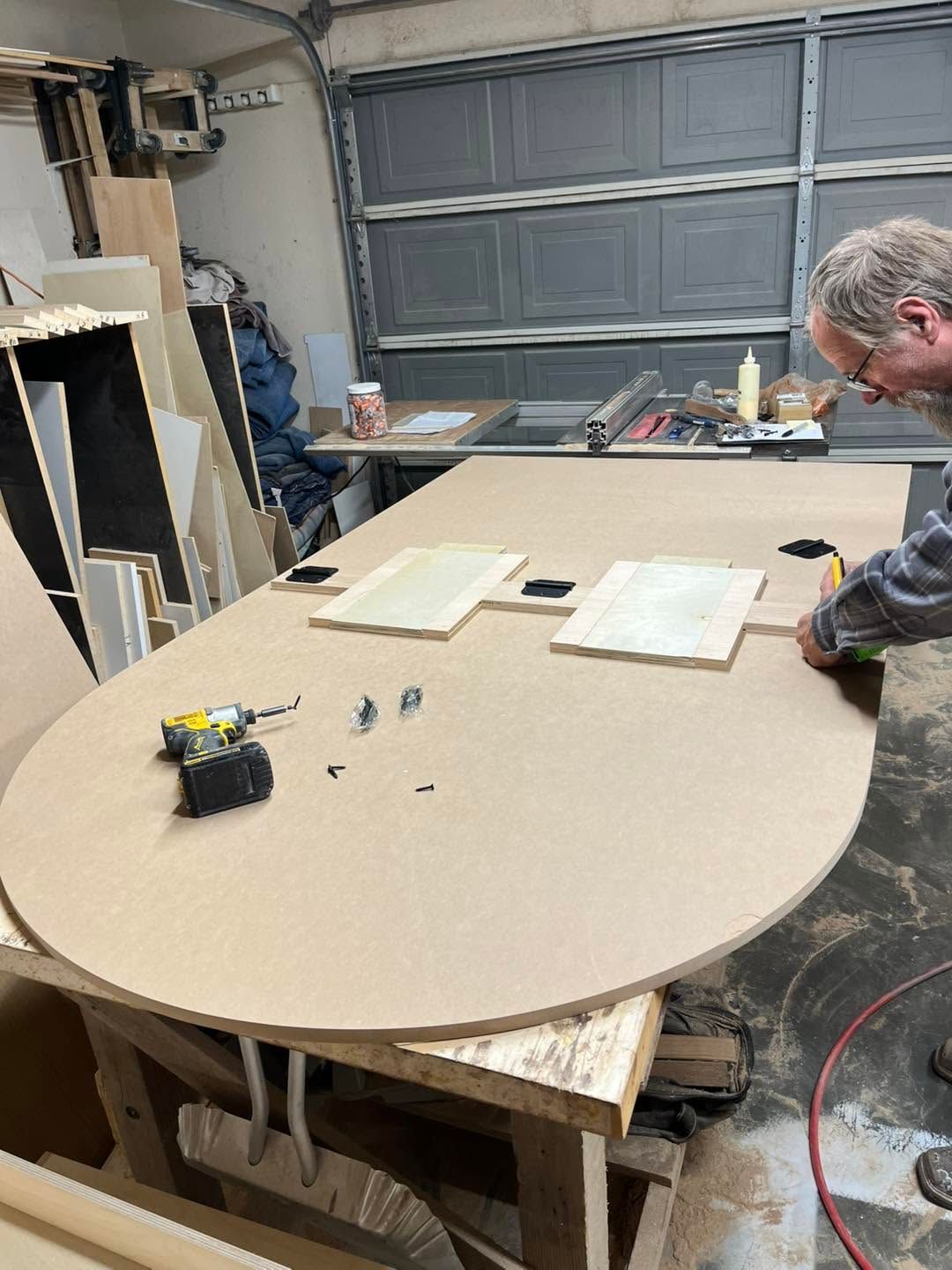 Person working on a large oval tabletop in a garage workshop with tools and materials spread out.