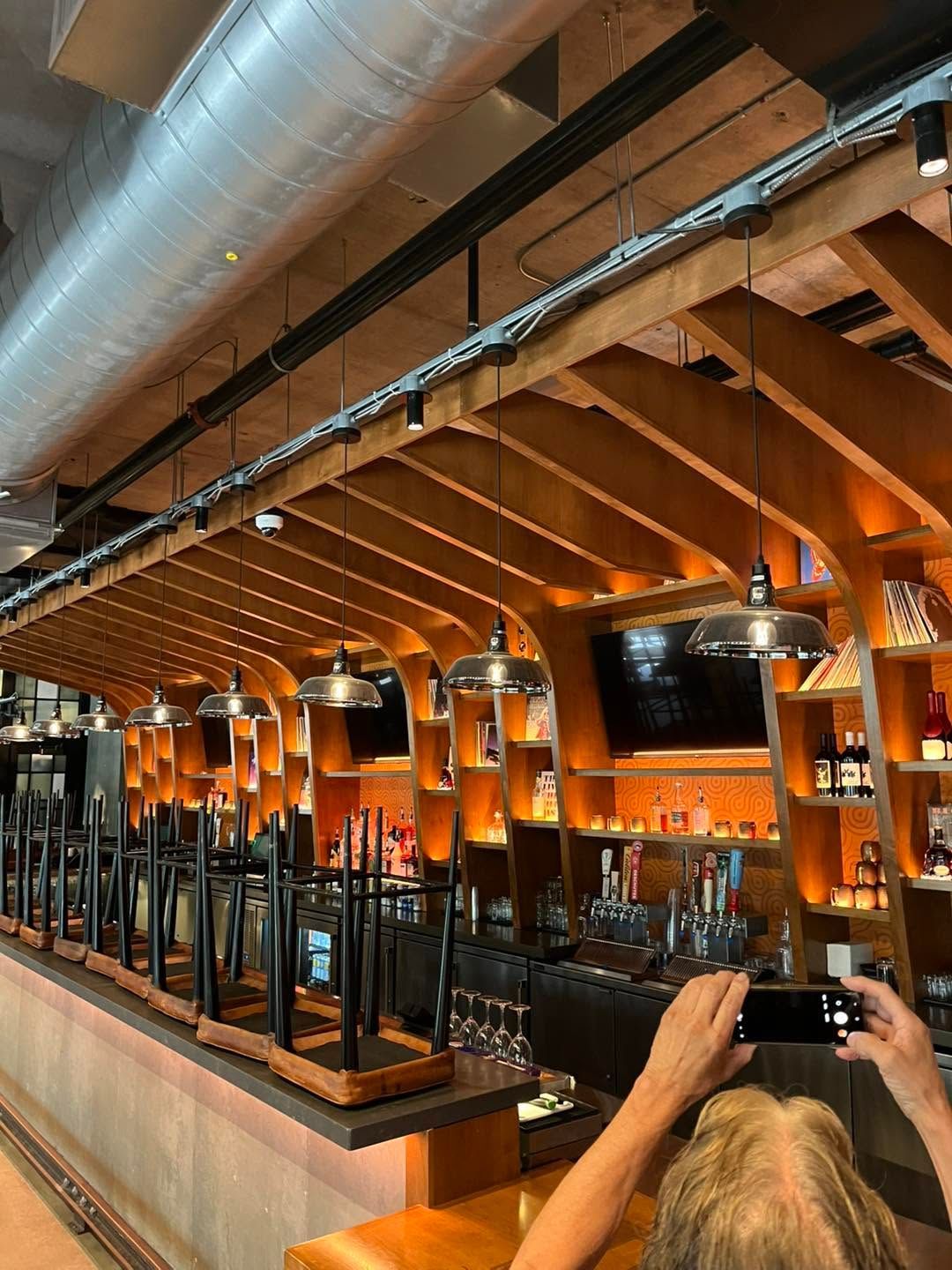 Modern restaurant interior with exposed wood beams, orange lighting, and diners seated at tables.