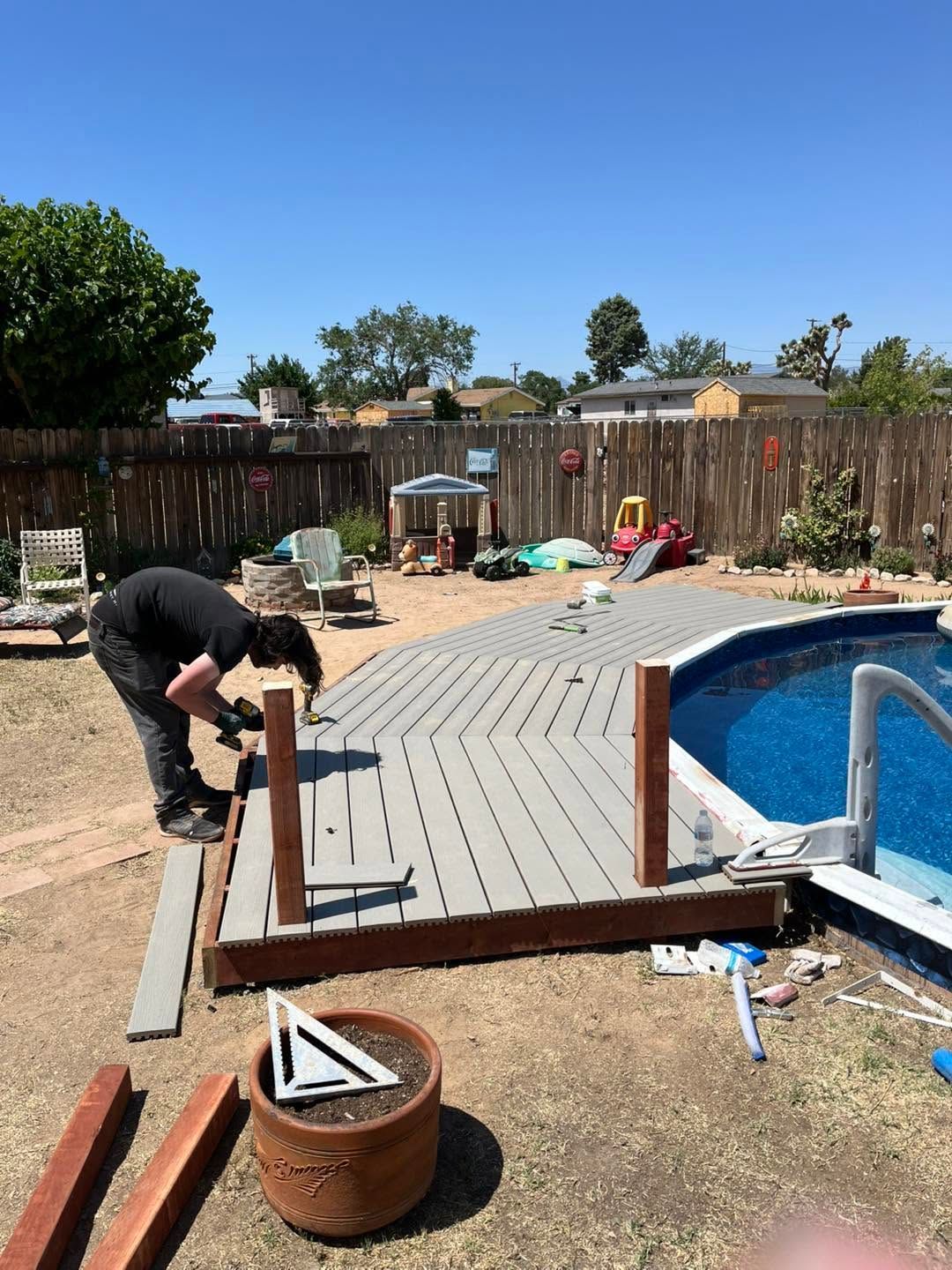 Person building a gray deck beside a blue pool in a sunny backyard