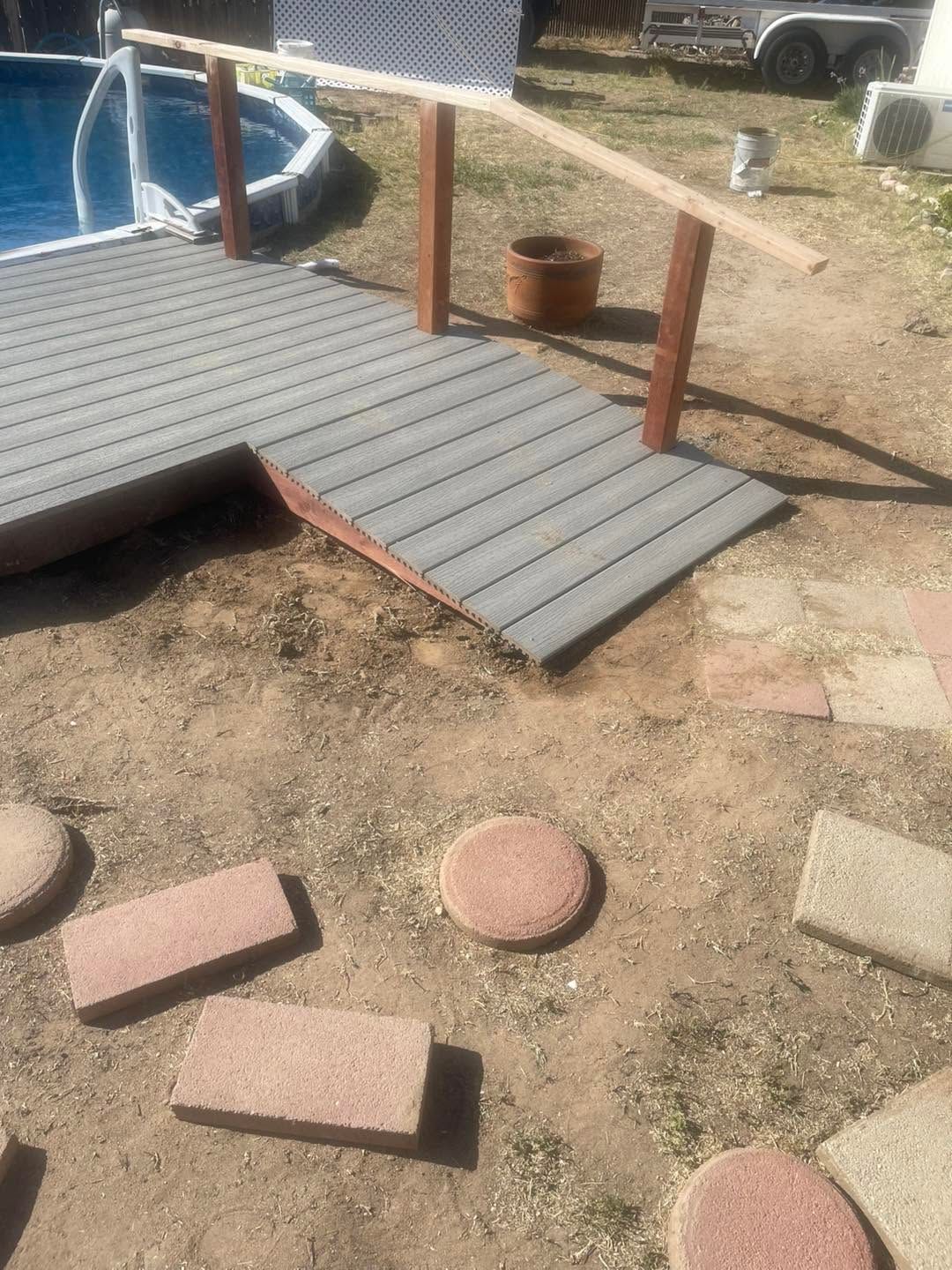 Small wooden ramp and steps leading to a raised deck in a dirt yard with potted plants nearby.