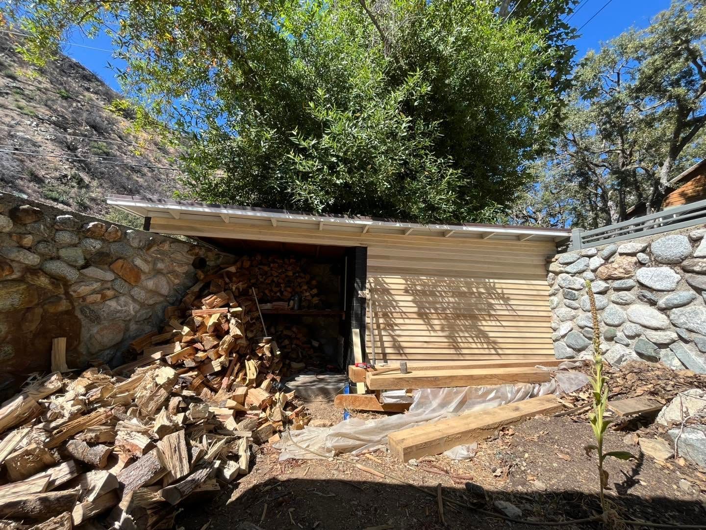 Small shed with stacked firewood, tan siding, and stone retaining walls in a wooded outdoor area.