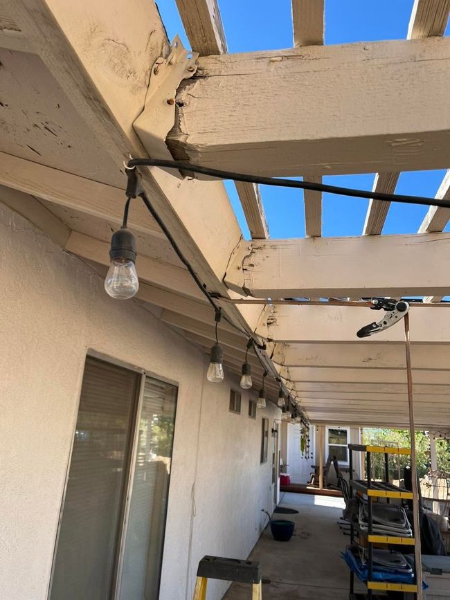 Covered outdoor walkway with exposed beams, hanging bulbs, and stacked items along a beige wall.