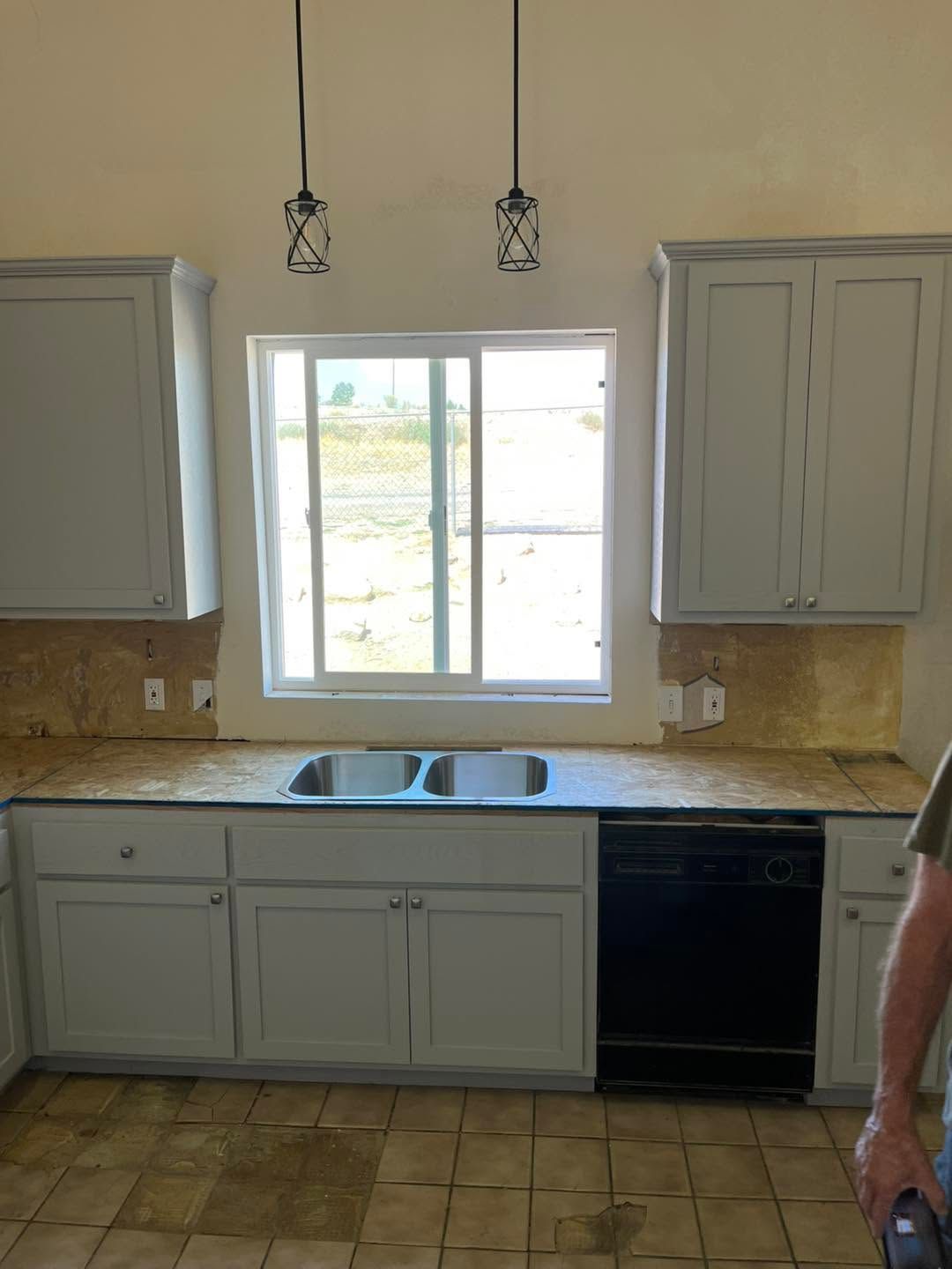 Kitchen with gray cabinets, white sink under a window, tan tile backsplash and floor, and pendant lights above.