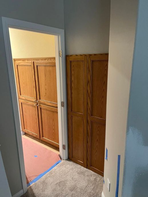 Hallway with open doorway to a room of wooden cabinets and a narrow closet; blue painter’s tape on the floor.