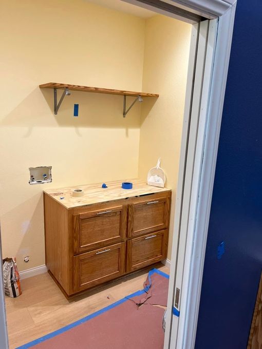 Small bathroom under renovation with a wooden vanity, shelf, and blue painter’s tape on the floor.