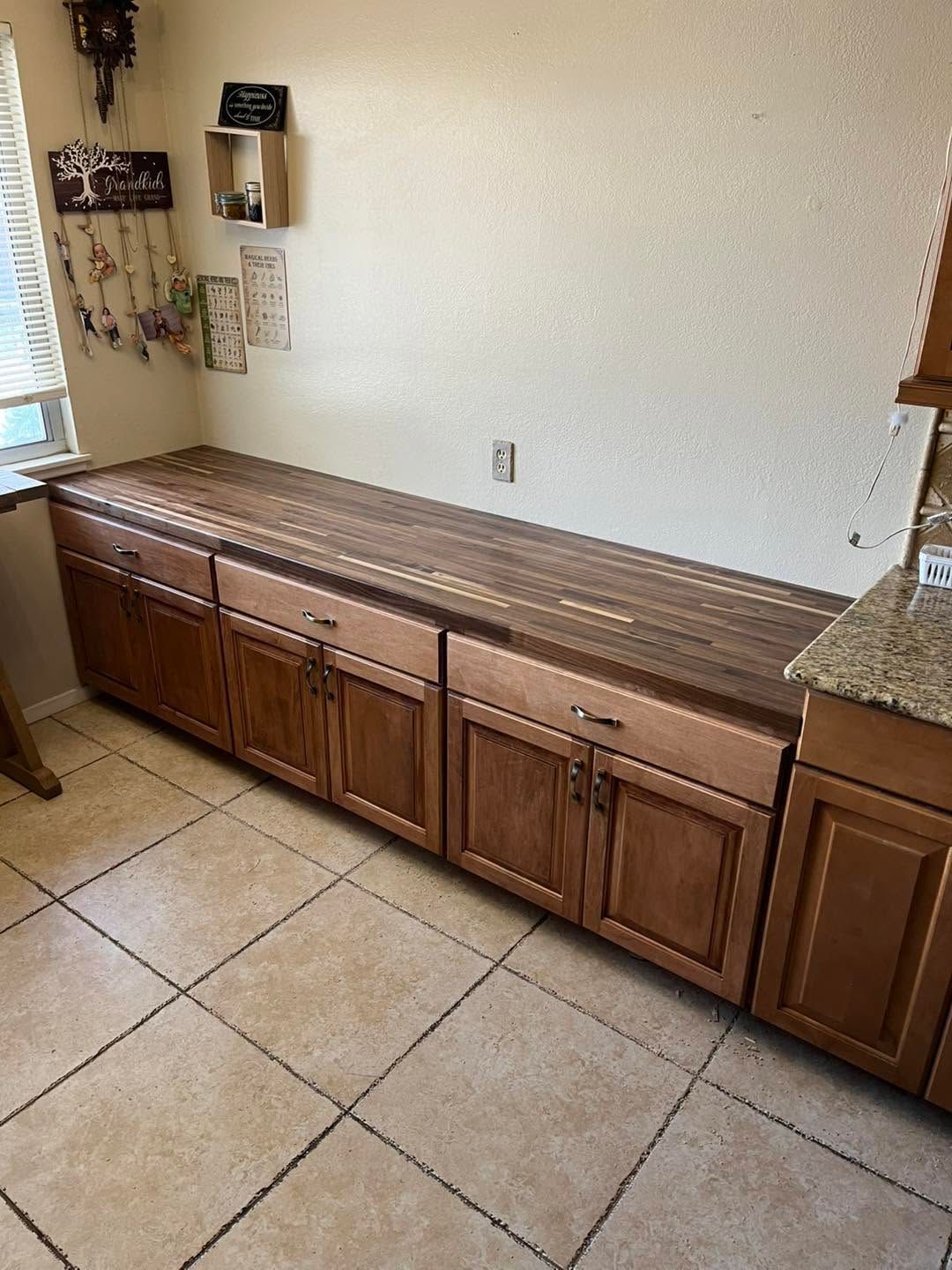 Kitchen with long wooden cabinets and countertop against a white wall and tiled floor.